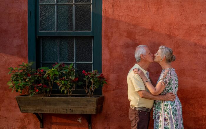Senior couple kissing lovingly against a quaint red wall with a window and plants.