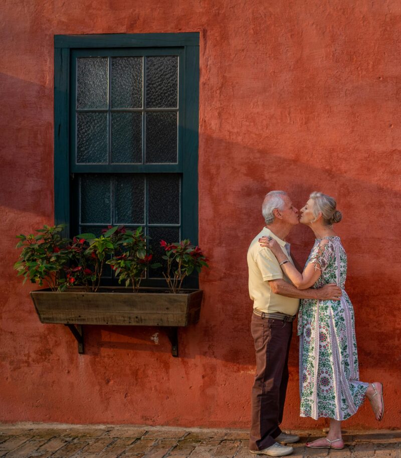 Senior couple kissing lovingly against a quaint red wall with a window and plants.