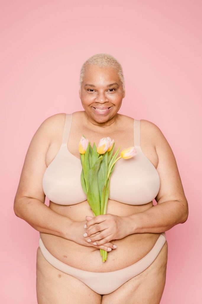 Happy plump African American female model in lingerie looking at camera while standing on pink background with flowers in studio